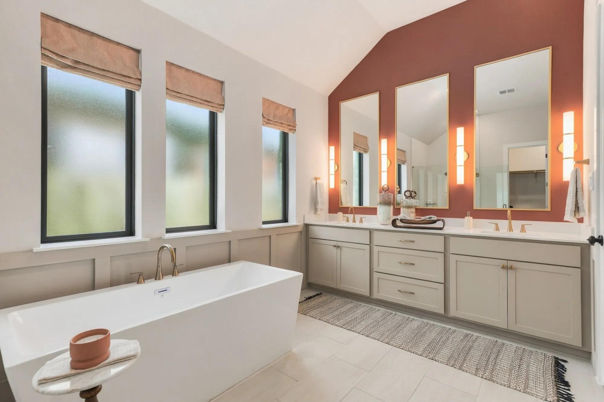 Sophisticated custom home primary bathroom with wainscoting behind freestanding tub and double sink vanity with gold sconce lighting and pink accent wall